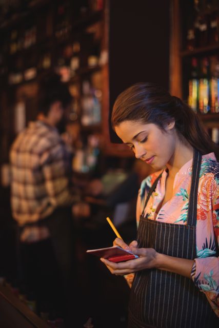 Waitress in a cozy pub taking an order on a notepad, wearing an apron. Ideal for themes related to hospitality, customer service, nightlife, and casual dining. Can be used for articles, blogs, or advertisements focusing on restaurant work, bar service, or the dining experience.