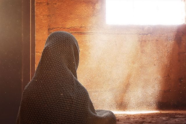 Person is sitting wrapped in a shawl in a dusty room illuminated by a shaft of sunlight from a window. This evokes feelings of solitude and contemplation. The warm sunlight and dusty air create a vintage, rustic atmosphere. The image can be used in contexts related to meditation, peaceful moments, introspection, spirituality, and artistic photography.