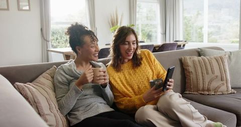 Female friends laughing and scrolling smartphone on cozy living room sofa with coffee