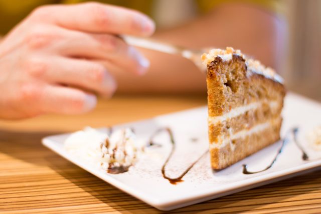 Person holding fork while enjoying a slice of carrot cake with cream frosting on a white plate. Ideal for use in food blogs, dessert recipes, bakery advertisements, and culinary magazines showing delicious snacks and desserts in casual dining settings.