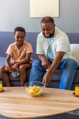 African american father and son gaming and sharing snacks on sofa at home - bonding moment