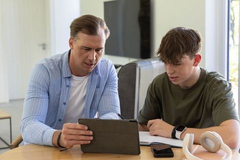 Father guiding teenage son using tablet and writing in notebook at home table
