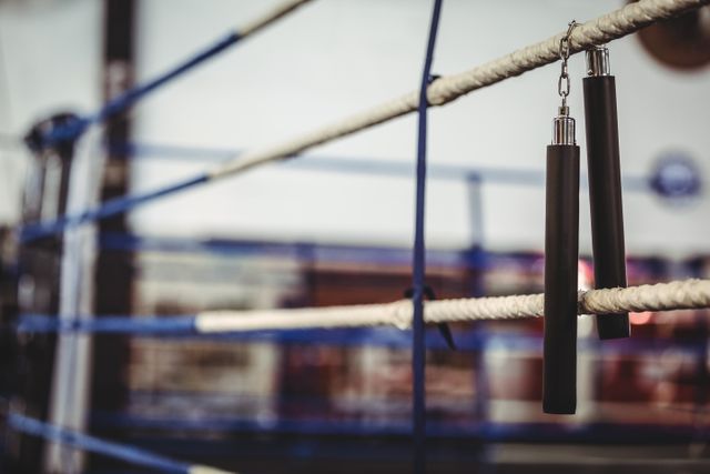 Nunchaku hanging on boxing ring ropes in a fitness studio. Ideal for use in articles or advertisements related to martial arts training, combat sports, gym equipment, or fitness routines. Can also be used in blogs or social media posts about self-defense techniques or traditional martial arts practices.