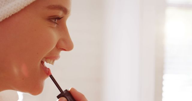 Young woman is applying lip gloss while smiling. Suitable for beauty, skincare or personal care campaigns, as well as content focusing on morning routines and healthy lifestyles.