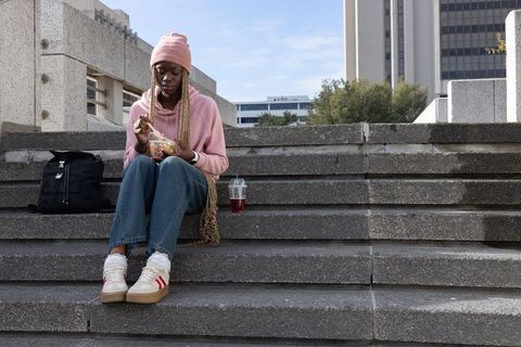 African american student sitting on concrete steps eating salad, wearing pink hoodie