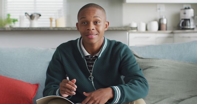 Teen boy sits on couch with notebook, appearing engaged in his studies in a cozy home environment. Ideal for use in educational materials, tutoring ads, and articles about student lifestyles.