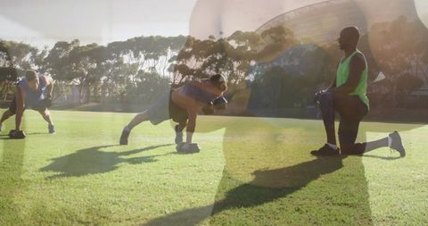 Outdoor group performing dumbbell rows on sunlit grass with coach kneeling guiding