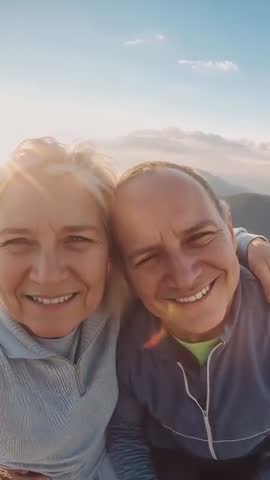 Vertical video of smiling senior couple taking selfie at mountain sunset with sun flare
