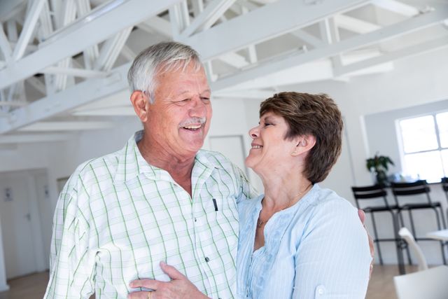 This image captures a joyful senior couple embracing and smiling indoors, showcasing their affectionate relationship. Ideal for use in advertisements, articles, or brochures related to senior living, retirement communities, healthy aging, and lifestyle content. The natural light and casual setting make it perfect for promoting family values, love, and happiness in older age.