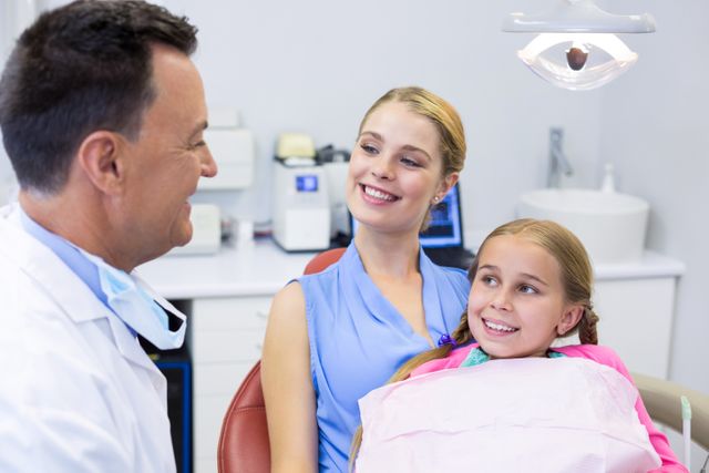 Dentist engaging with a young patient and her mother at a dental clinic. The child is sitting in the dental chair, smiling, while the mother looks on with a smile. This image can be used for promoting dental health services, pediatric dentistry, family dental care, and healthcare advertisements.