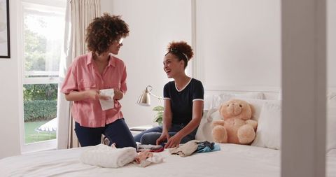 African American mother and daughter folding laundry and laughing in sunlit bedroom