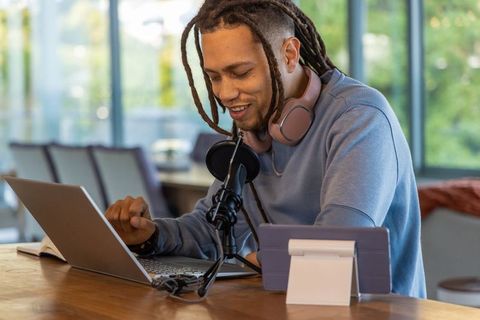African american creator podcasting on laptop with headphones and mic in sunlit cafe