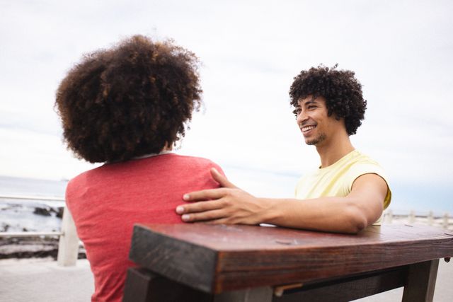 Happy afro african american young couple spending quality time while sitting on bench at beach. unaltered, lifestyle, love and togetherness concept.