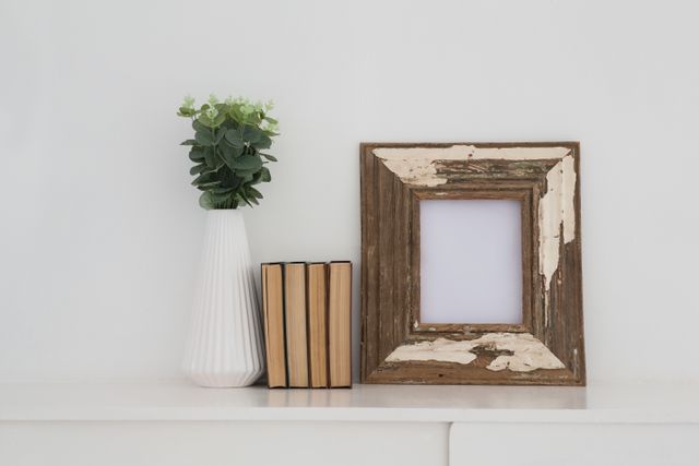 Rustic wooden frame with distressed finish, white vase with green plant, and stack of books arranged on white table. Ideal for home decor inspiration, minimalist interior design concepts, and modern living spaces. Perfect for blogs, magazines, and websites focused on home styling and decoration.