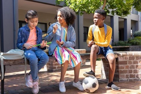 Diverse school children sitting on bench sharing snack and soccer ball at outdoor break