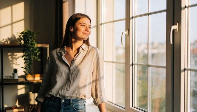 Woman Standing by Sunlit Window Wearing Linen Shirt and Jeans, Golden Hour Calm