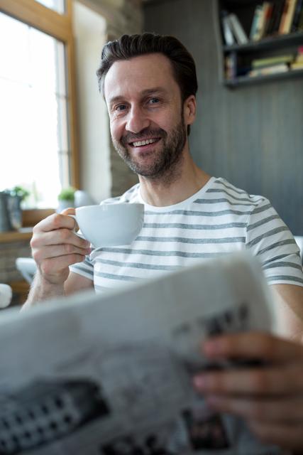 Smiling man holding a cup of coffee and reading newspaper in coffee shop