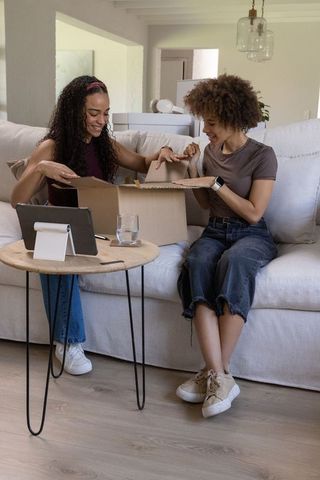 African american women unpacking shipping box and checking tablet on sofa in modern minimalist home