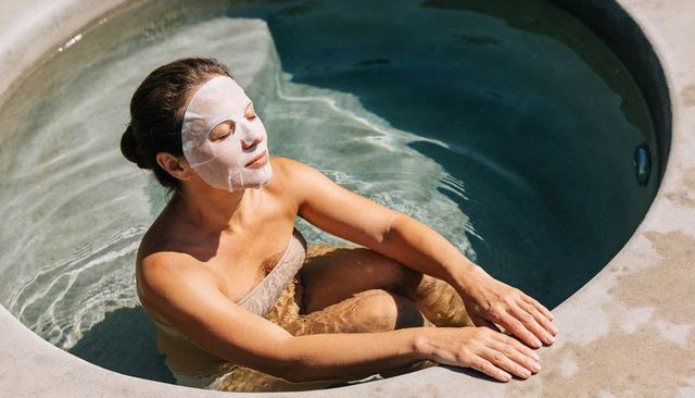 Woman soaking in outdoor spa tub wearing sheet mask and towel, relaxing wellness moment