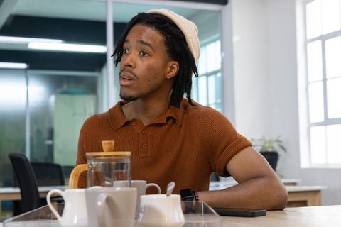 African american man pouring tea in coworking office wearing knit hat rust polo, daylight