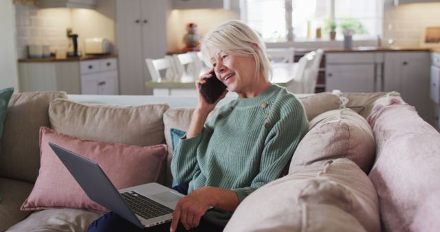 Senior woman sitting on cozy sofa in living room, working on laptop and talking on phone. Ideal for illustrating concepts of remote work, senior independent lifestyle, technology adoption by older adults, and modern home-based employment.
