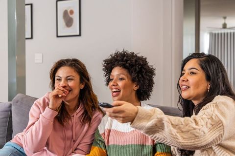 Laughing multicultural friends sitting on sofa watching tv and sharing remote in living room