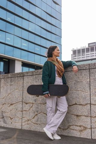 Urban female skater leaning on stone wall holding skateboard near reflective glass office building