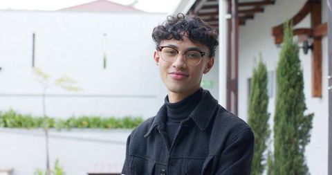 Young asian man standing and posing in courtyard wearing black jacket and round glasses