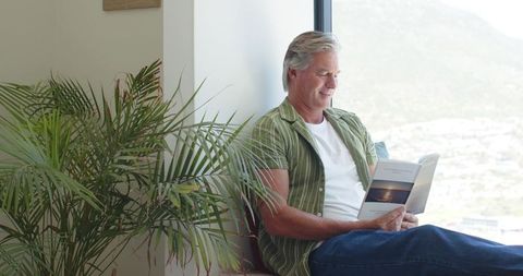 Senior man reading paperback by sunlit window with indoor palm, relaxed home leisure lifestyle
