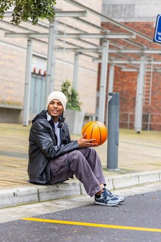 Urban young man holding basketball sitting on curb in beanie leather jacket and headphones