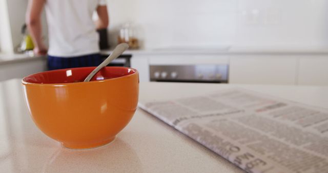 This image showcases a modern kitchen with a focus on the daily morning routine. The foreground features an orange cereal bowl and a folded newspaper on a white countertop, while a person is seen in the background, possibly preparing breakfast. Ideal for illustrating themes of domestic life, morning rituals, healthy eating, or kitchen designs in lifestyle blogs, advertisements, or home décor magazines.