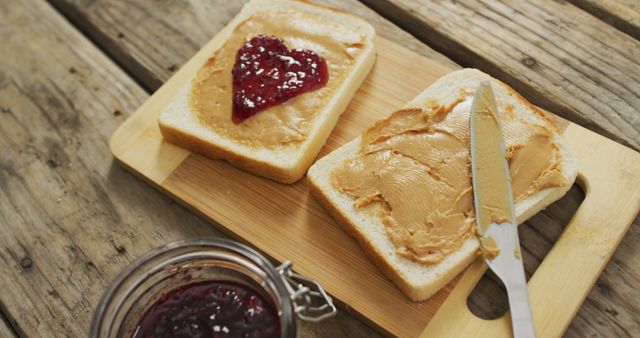 This image shows two slices of bread on a wooden cutting board. One slice has a spread of peanut butter topped with a heart-shaped jelly. The other slice is spread with peanut butter. Next to the bread is a jar of jelly with a closed lid, and a butter knife rests on the cutting board. Ideal for use in articles or ads related to breakfast, snacks, food preparation, or themes of love and care.