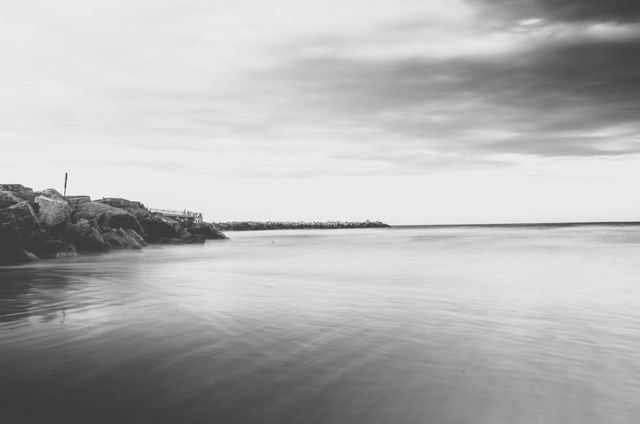 This tranquil seascape in black and white features a rocky shoreline and calm water under an overcast sky. The minimalist and peaceful atmosphere makes it suitable for use in designs or decor emphasizing serenity and simplicity. Ideal for background images, marketing materials for travel or nature experiences, and as calming wall art in homes or office spaces.
