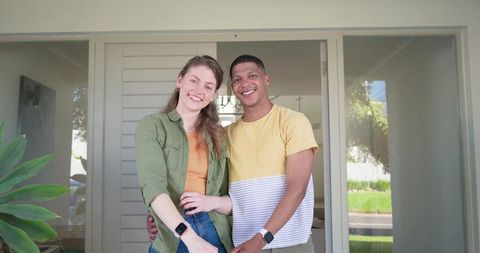 Smiling mixed-race couple standing at front door wearing smartwatches and casual clothes
