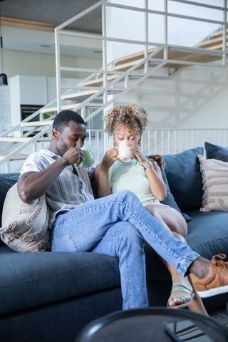 Diverse couple sipping coffee on navy sofa, relaxing in modern minimalist living room