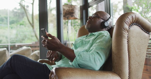 Senior man relaxing in a comfortable chair with headphones on, listening to music or a podcast on his smartphone. Ideal for topics related to technology use in older adults, promoting relaxation and leisure activities, and advertising living room furniture.