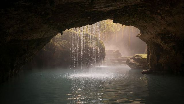 Sunlight streaming through sinkhole onto cascading water curtains over mossy boulders