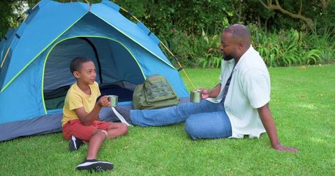 African american father and son sharing camping mugs at tent entrance, bonding outdoors