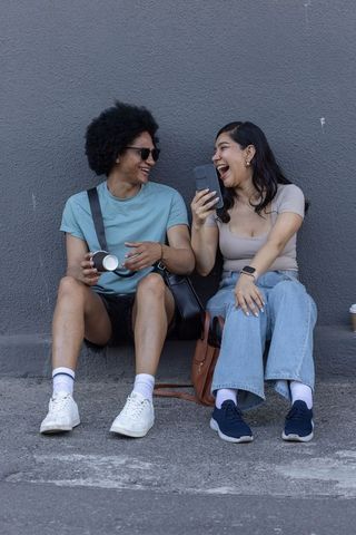 Diverse friends laughing and sharing smartphone while sitting on urban curb holding coffee