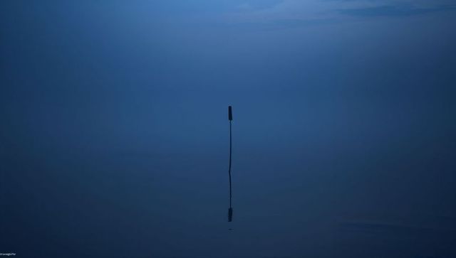 Minimalist twilight seascape with single slender post reflecting on glassy calm water
