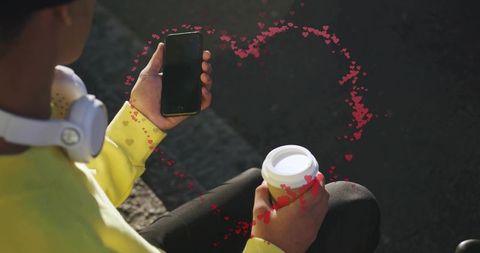 Urban young man using smartphone and holding takeaway coffee with heart overlay on street