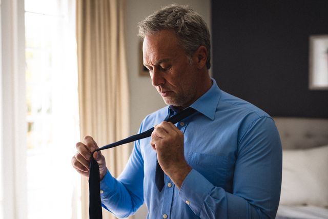 Mature man tying a tie in a bedroom, wearing a blue shirt. Ideal for themes related to morning routines, getting ready for work, lifestyle, and home life. Can be used in articles or advertisements focusing on business attire, preparation, and daily routines.