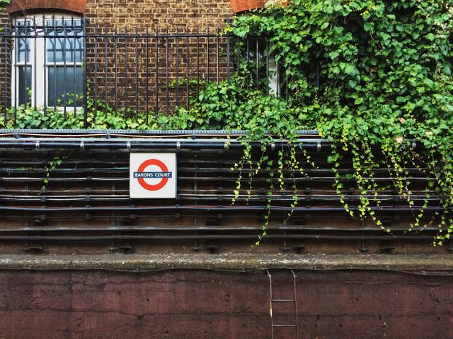 Classic London Underground station seen from track level with a Barons Court sign and creeping ivy adding an urban touch. This photograph might be used for travel websites, transportation services, urban gardening, or showcasing London's mix of nature and city life.