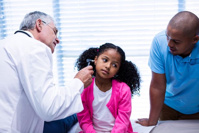 Doctor examining young girl's ear with otoscope while father watches. Useful for topics related to pediatric healthcare, medical consultations, family health, and doctor-patient interactions.
