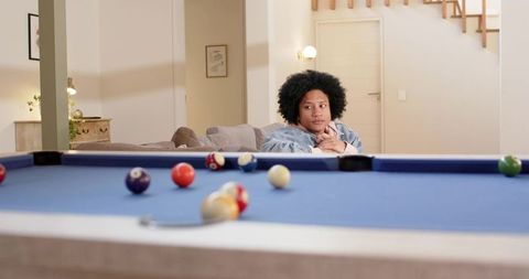 Contemplative african american woman leaning on blue-felt pool table in cozy living room