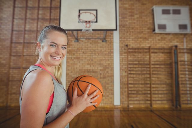High school girl smiling while holding a basketball in a gymnasium. Ideal for use in educational materials, sports promotions, youth fitness campaigns, and school-related content.