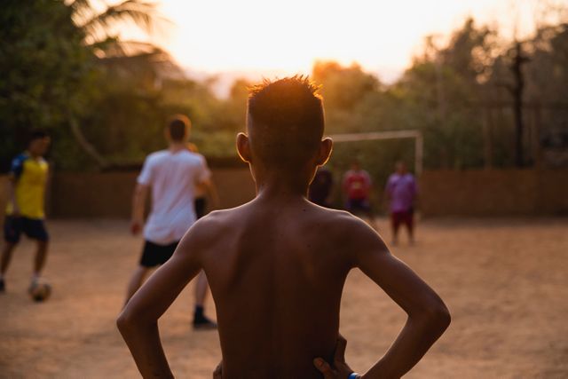 Young boys engaging in a soccer match on a dirt field at sunset. Seen from behind, with one boy standing in the foreground. Shirtless and deeply involved in game, capturing energy and freedom of childhood. Suitable for themes of youth sports, team activities, outdoor play, community, and summer evenings.
