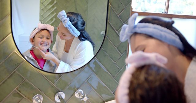 Mother and daughter in bathroom enjoying morning skincare routine. Both have headbands and are engaged in a joyful moment. Ideal for use in parenting blogs, family wellness ads, beauty product promotions, and family-oriented campaigns focusing on morning routines, bonding, and skincare habits.