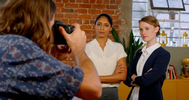 Two professional women standing with arms crossed, posing for a photo shoot in a modern office with exposed brick wall and large windows. A photographer is capturing their image. Useful for business promotions, teamwork representation, office culture themes, and corporate website content.