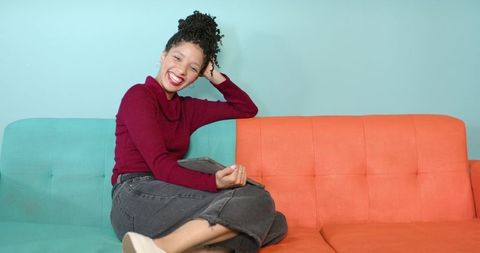 Smiling african american woman sitting on colorful two-tone sofa holding tablet, lifestyle